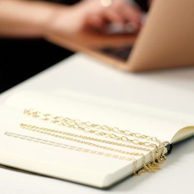 A notebook is open with three gold chains lying on it. In the background, a jewellery copywriter types product descriptions on a laptop.